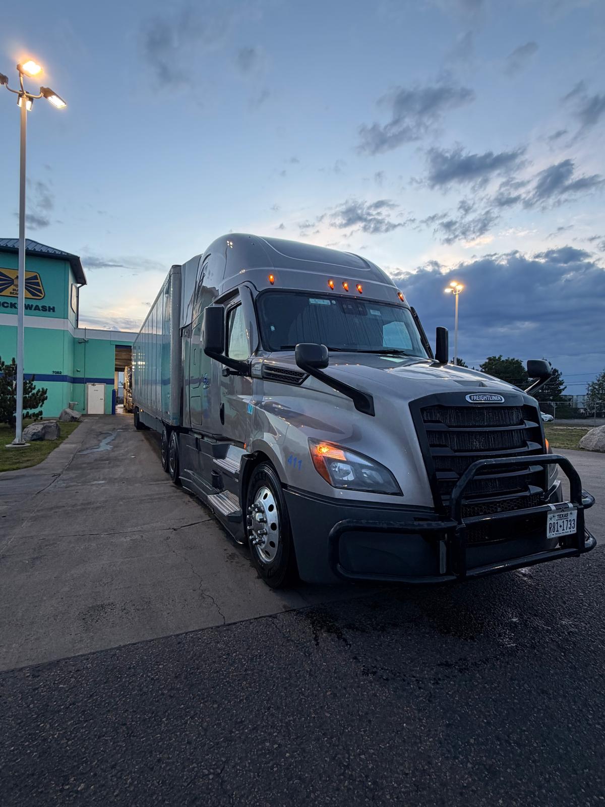 Silver Freightliner sleeper tractor with a trailer parked on pavement at dusk.